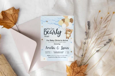 A baby shower invitation card with a brown teddy bear and balloons, placed beside an open beige envelope on a white fabric surface, accompanied by dried leaves and floral elements.